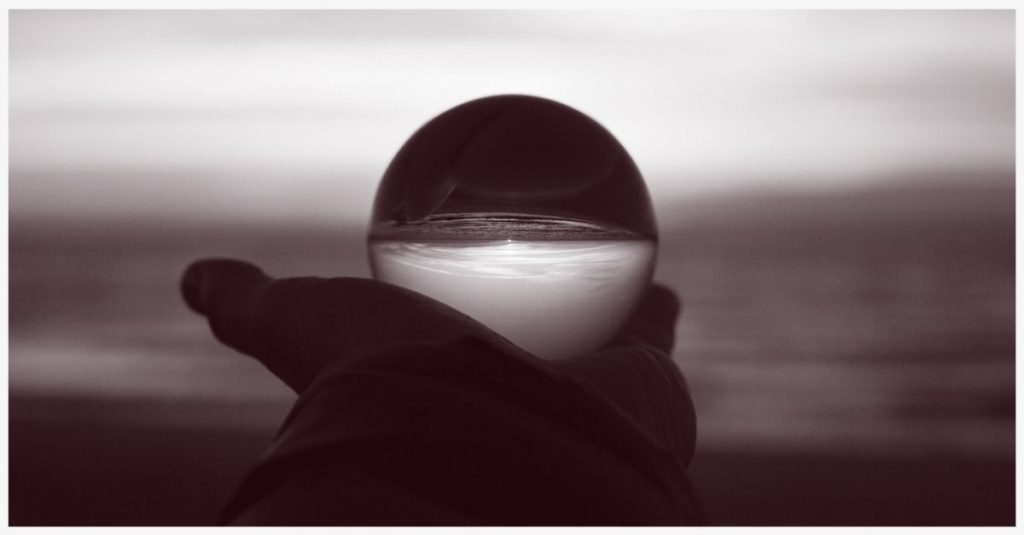 A glass orb held in hand with the image of an ocean sunset refracted through it.