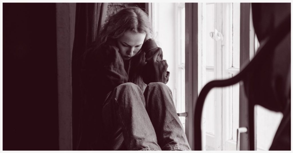 A young woman sitting next to window while holding her head in her hands