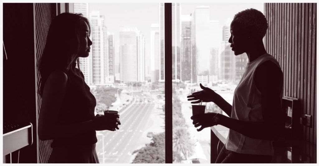 Two women talking in front of a window with a view of a city